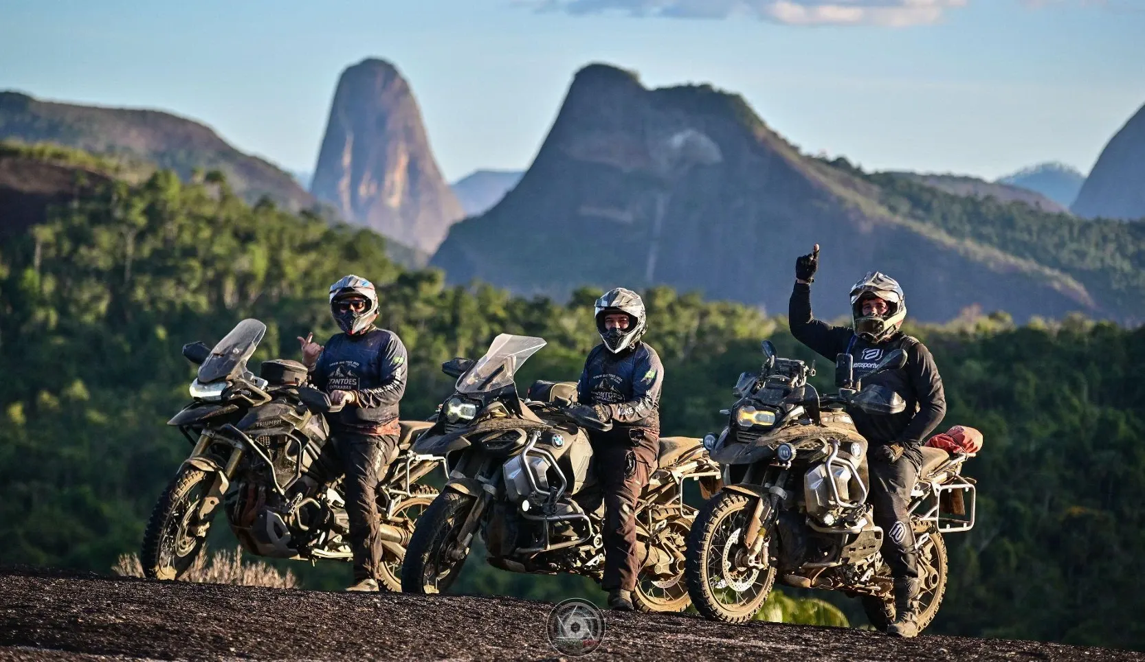 Três pilotos com motos big trail enlameadas sobre uma rocha, diante dos pontões de pedra e da mata
