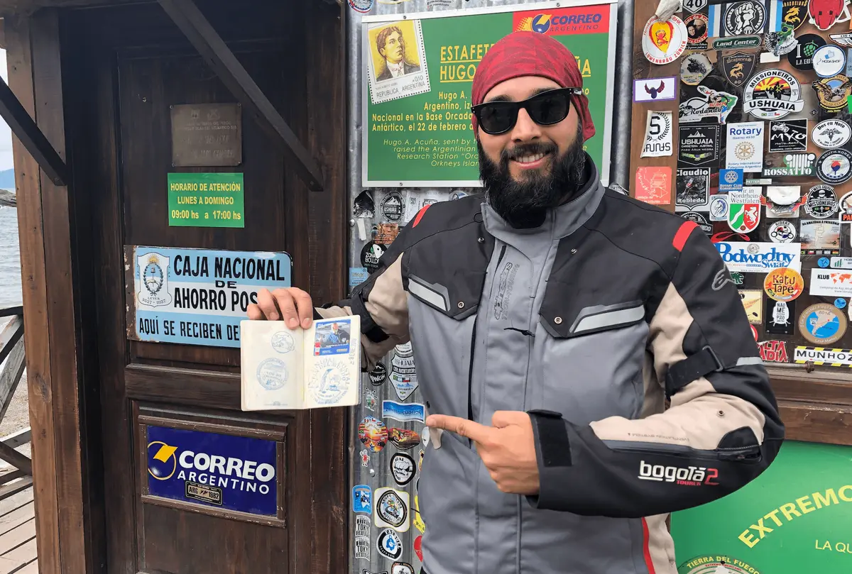 Thiago Berardi com jaqueta de moto segurando o passaporte em frente a um posto de fronteira